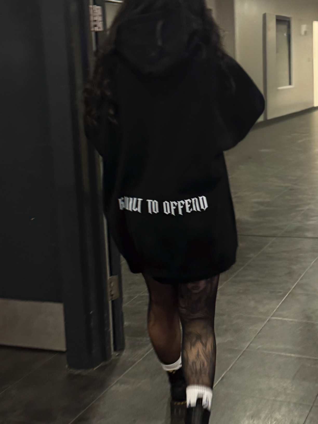Person wearing a black hoodie with text, standing in a dimly lit hallway.
